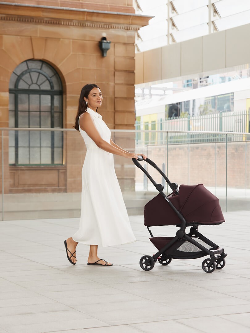 Close-up of the maroon Redsbaby SKIP³ Travel Pram being pushed by a woman in a white dress at the shops.