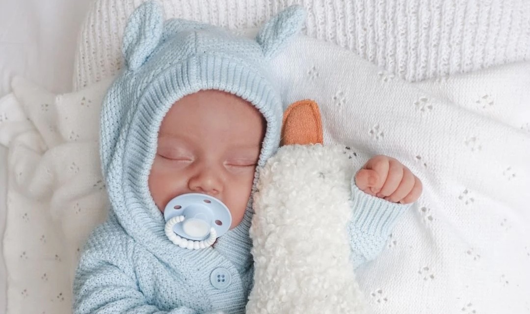 A baby in a blue teddy outfit asleep holding a toy duck with a CMC Gold Australian dummy in