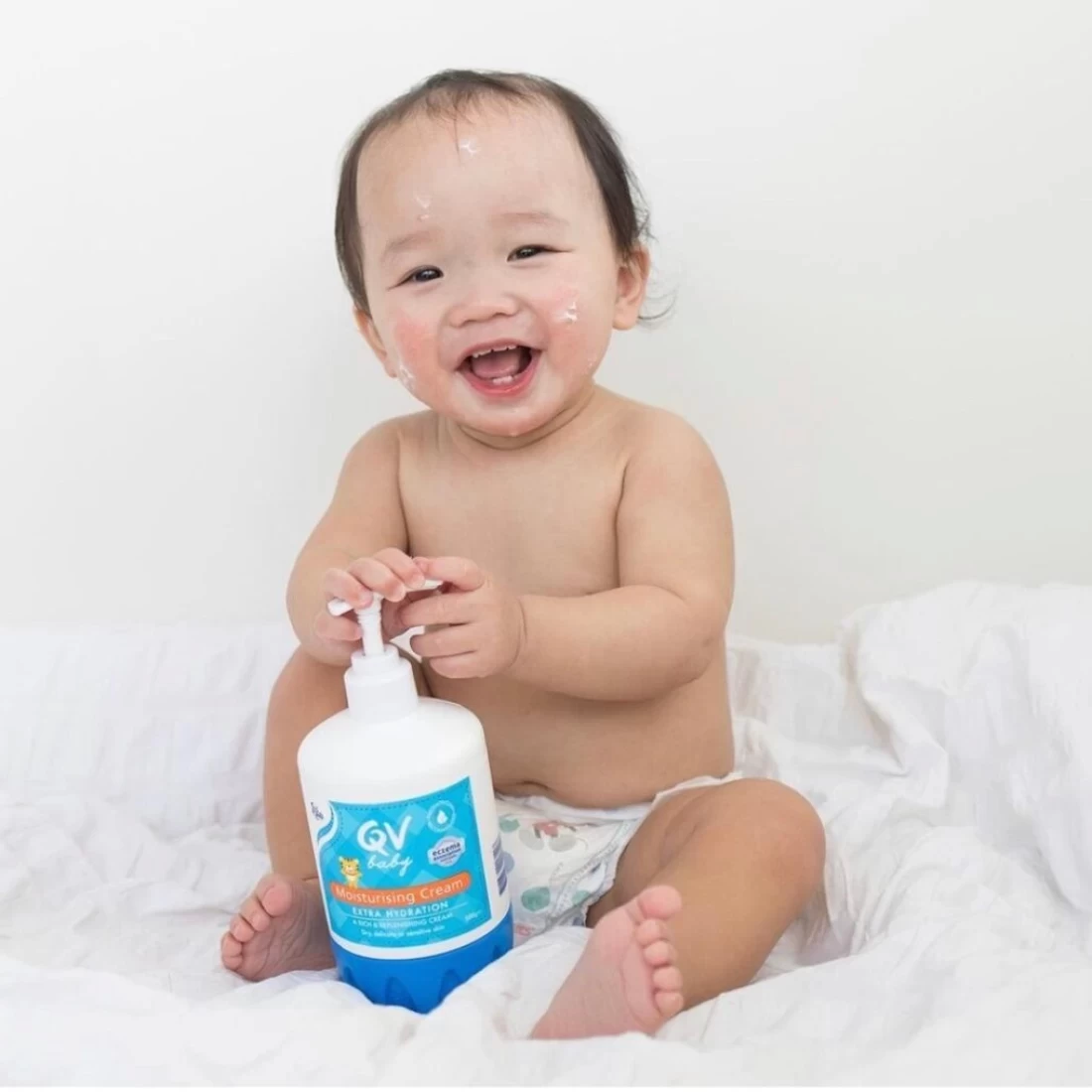 Newly bathed baby sitting on soft bed with gentle QV Baby Skincare product