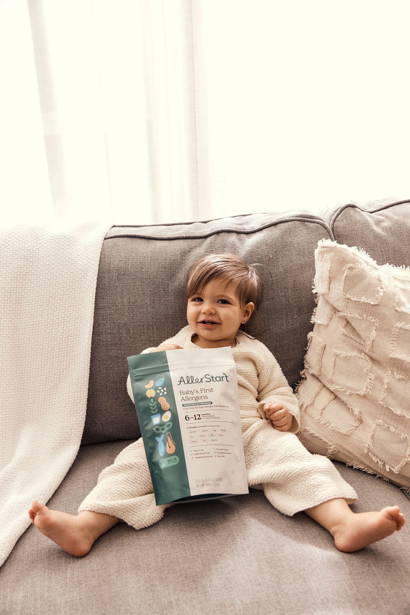 A baby seated on a couch while posing, holding baby food pouches. Best tips to starting solid food