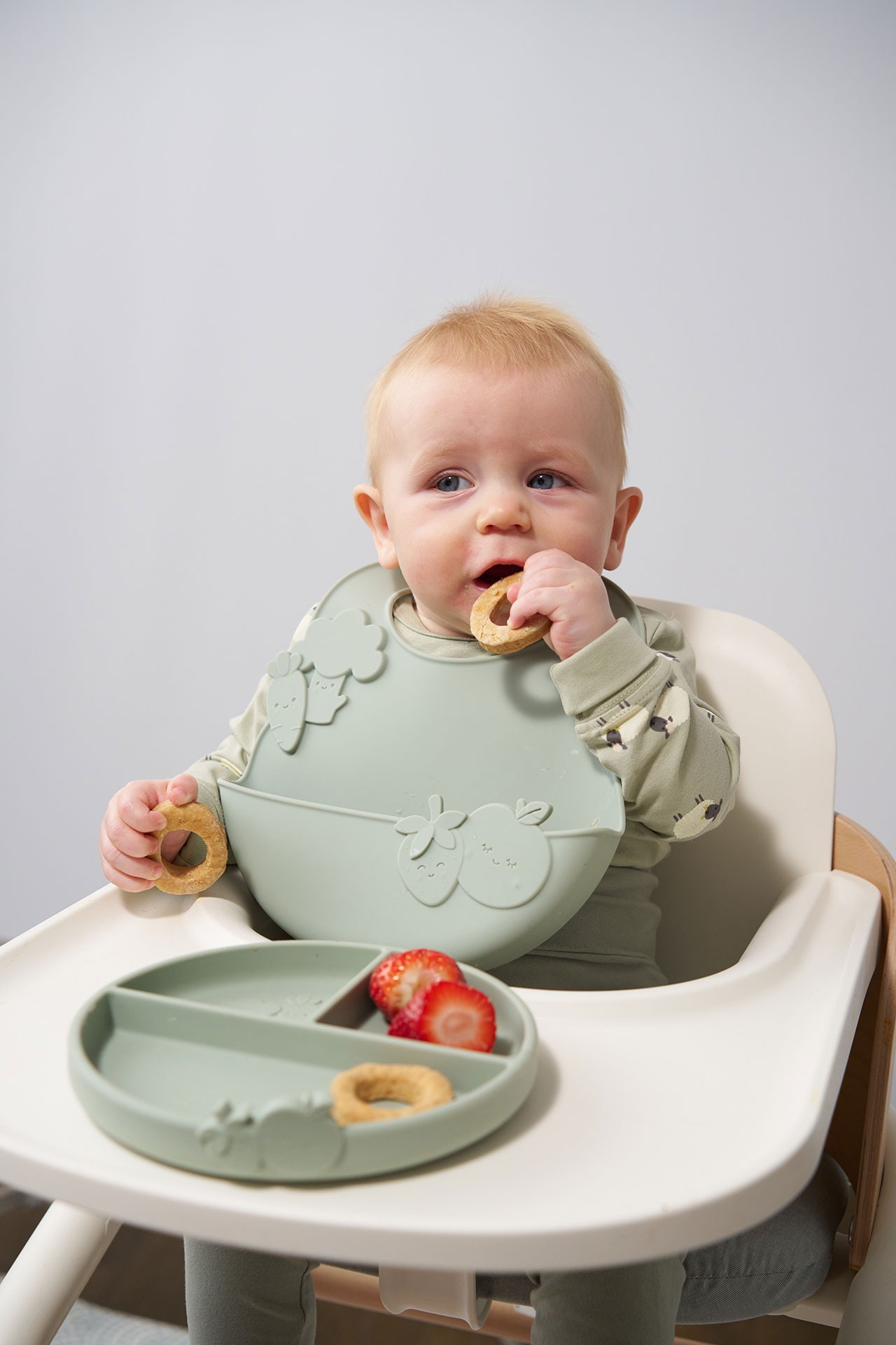 Baby seated in a high chair eating strawberries and cookies with age appropriate textures during baby led weaning.