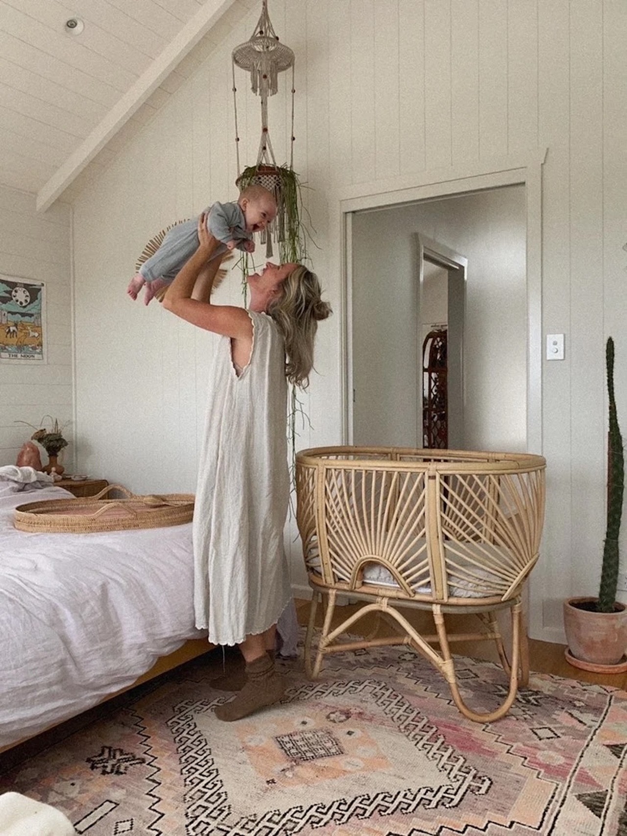 The Raja Homewares baby bassinet in a bedroom with a mum standing next to it holding her baby in the air and laughing