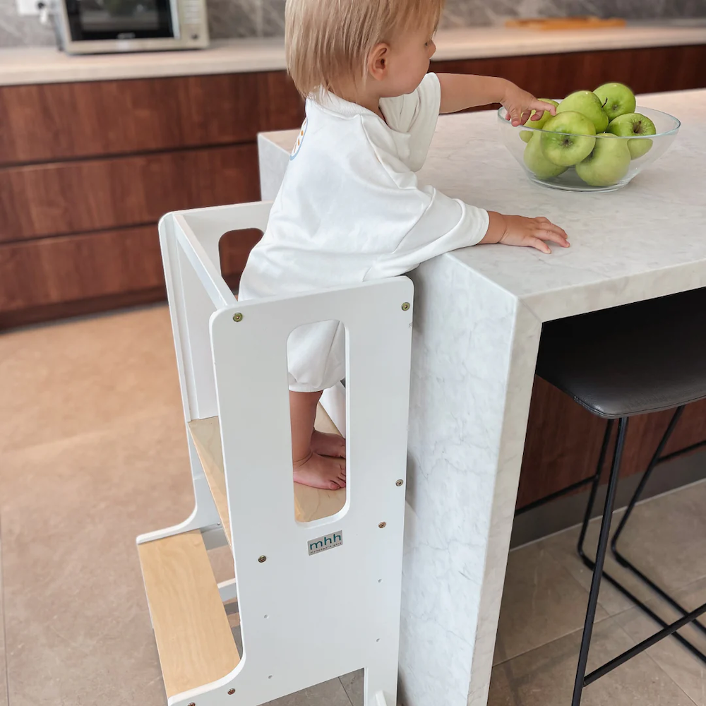 A toddler standing at a learning tower reaching for apples