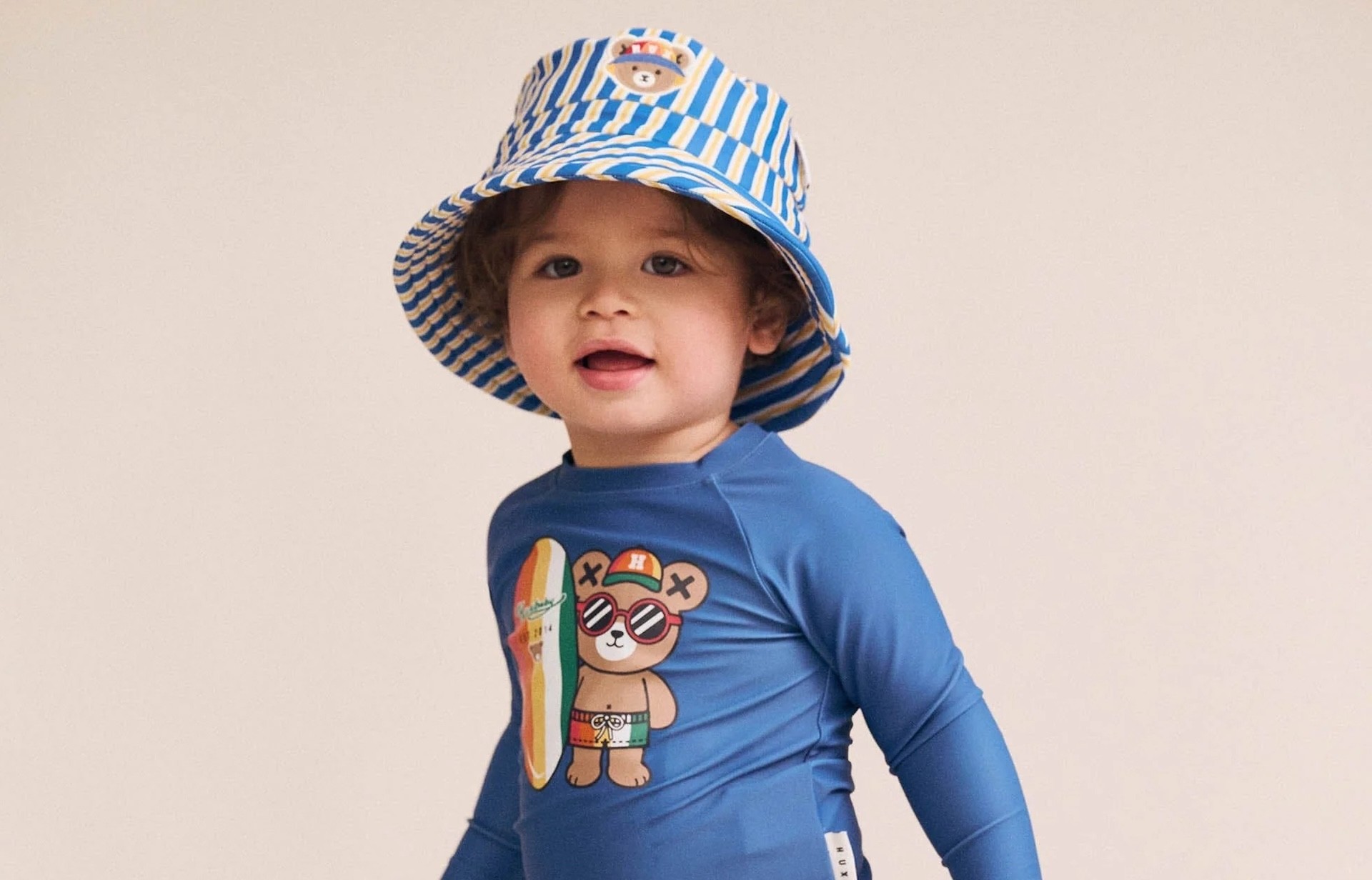 A baby in a striped beach hat and kids swimwear by Huxbaby