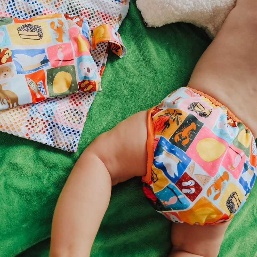 a baby wearing a colourful swim nappy, laying down on a beach towel