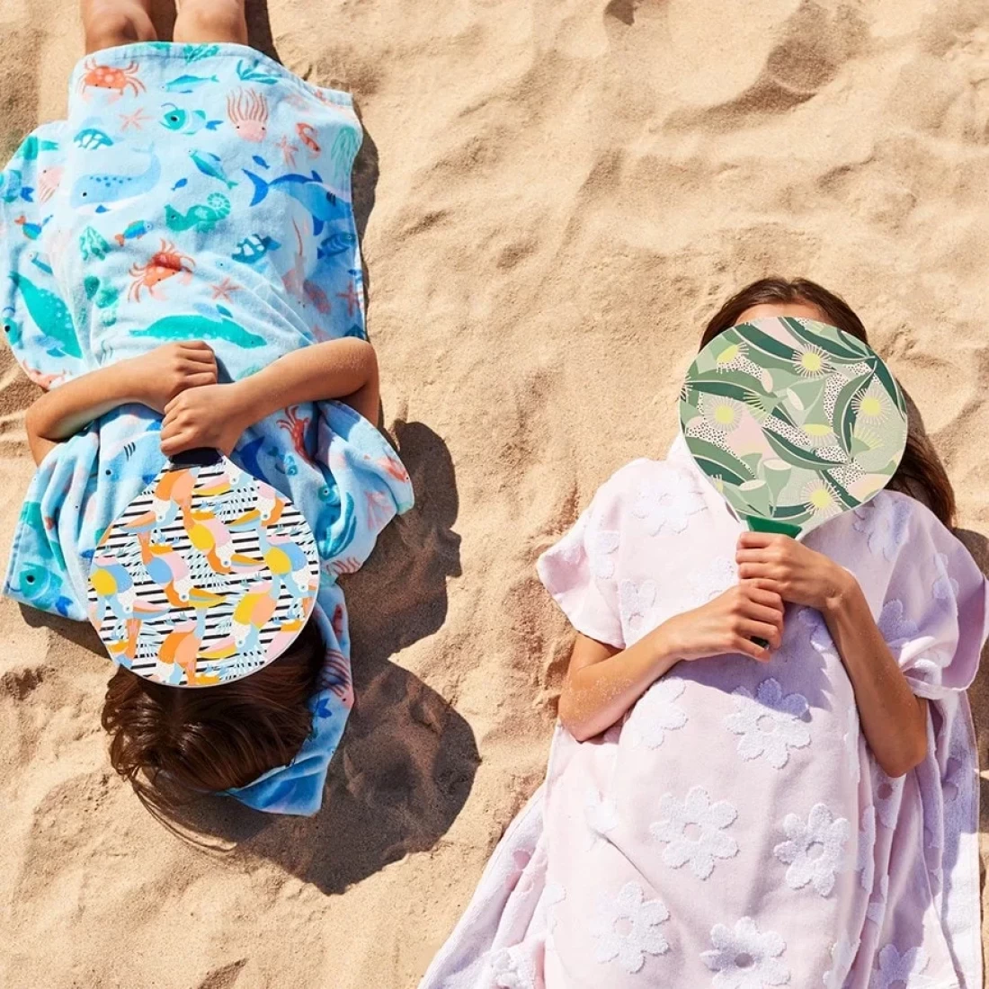 Two kids lying on the sand wearing the best hooded beach towel, perfect for beach days.