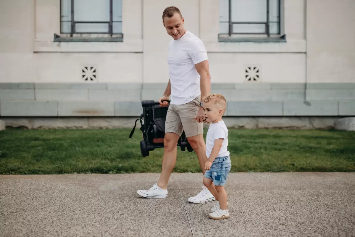 Dad carrying the folded Edwards & Co Otto – the best travel pram, with his toddler walking alongside him.