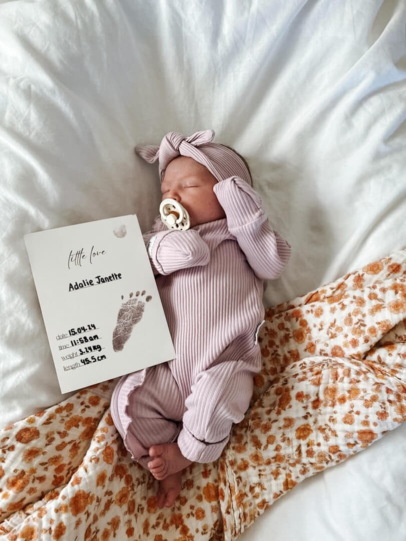 A sleeping  newborn baby wearing matching onesies and headband with an inkless milestone card stating baby's birthdate,, time, height, and weight