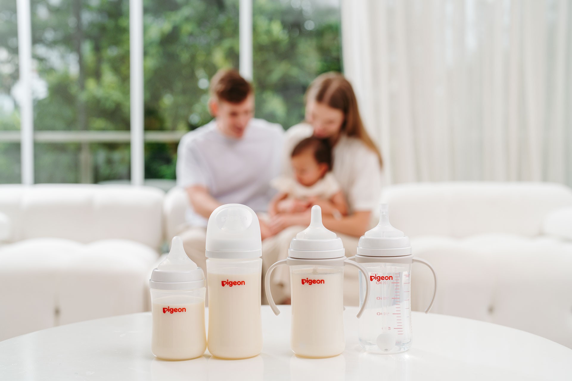 Pigeon baby bottles displayed in four different sizes on a table, with a mum, dad and baby bonding in the background