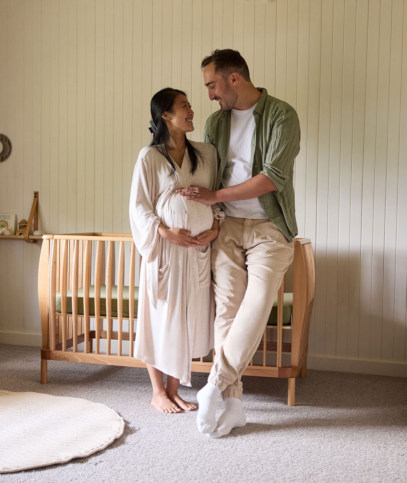Pregnant woman and partner standing by a wooden baby cot in a nursery, holding her belly bump and smiling at each other, choosing the best baby cot for their baby