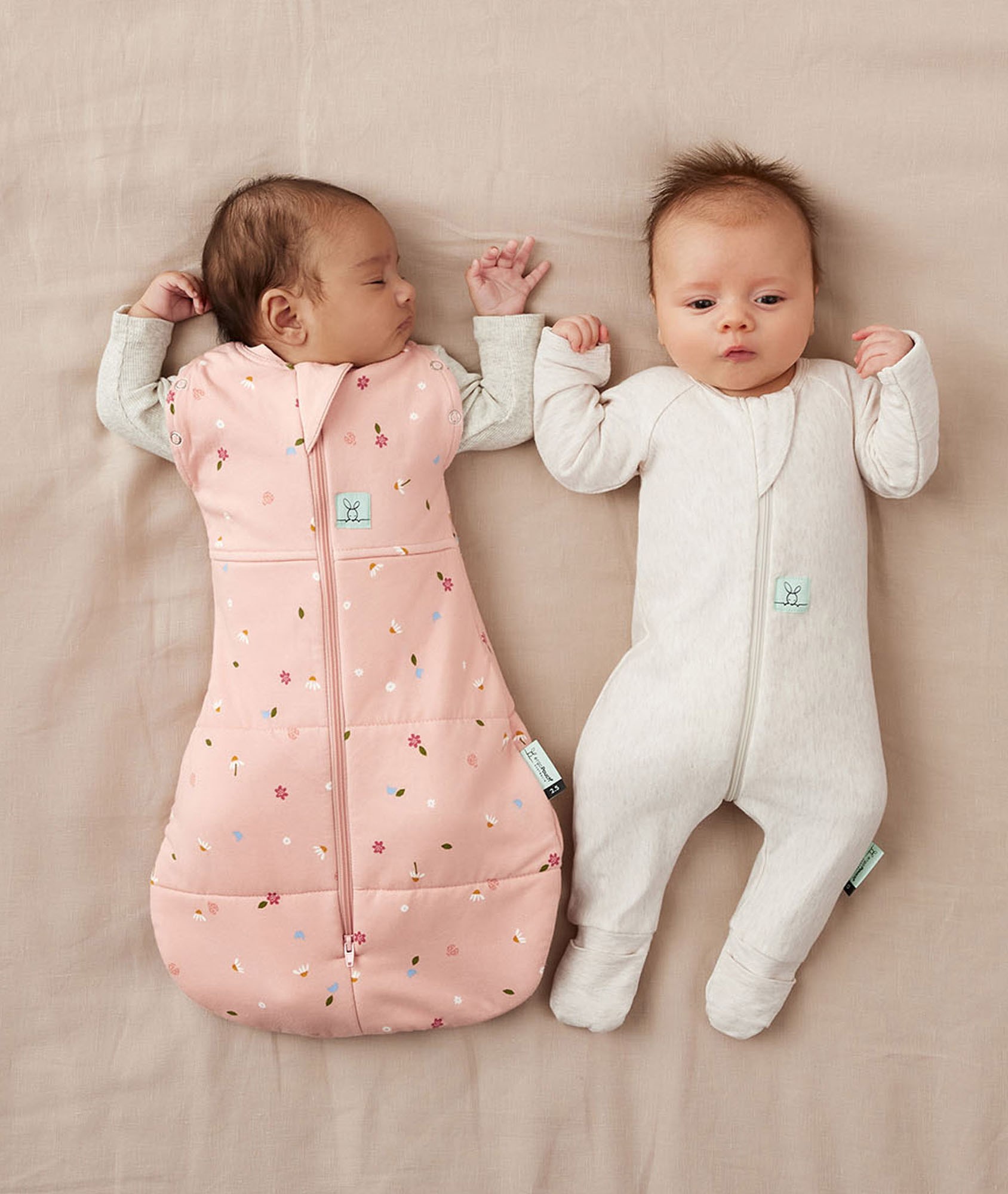 Newborn twins lying side by side in pink baby swaddle and white baby sleepsuits.