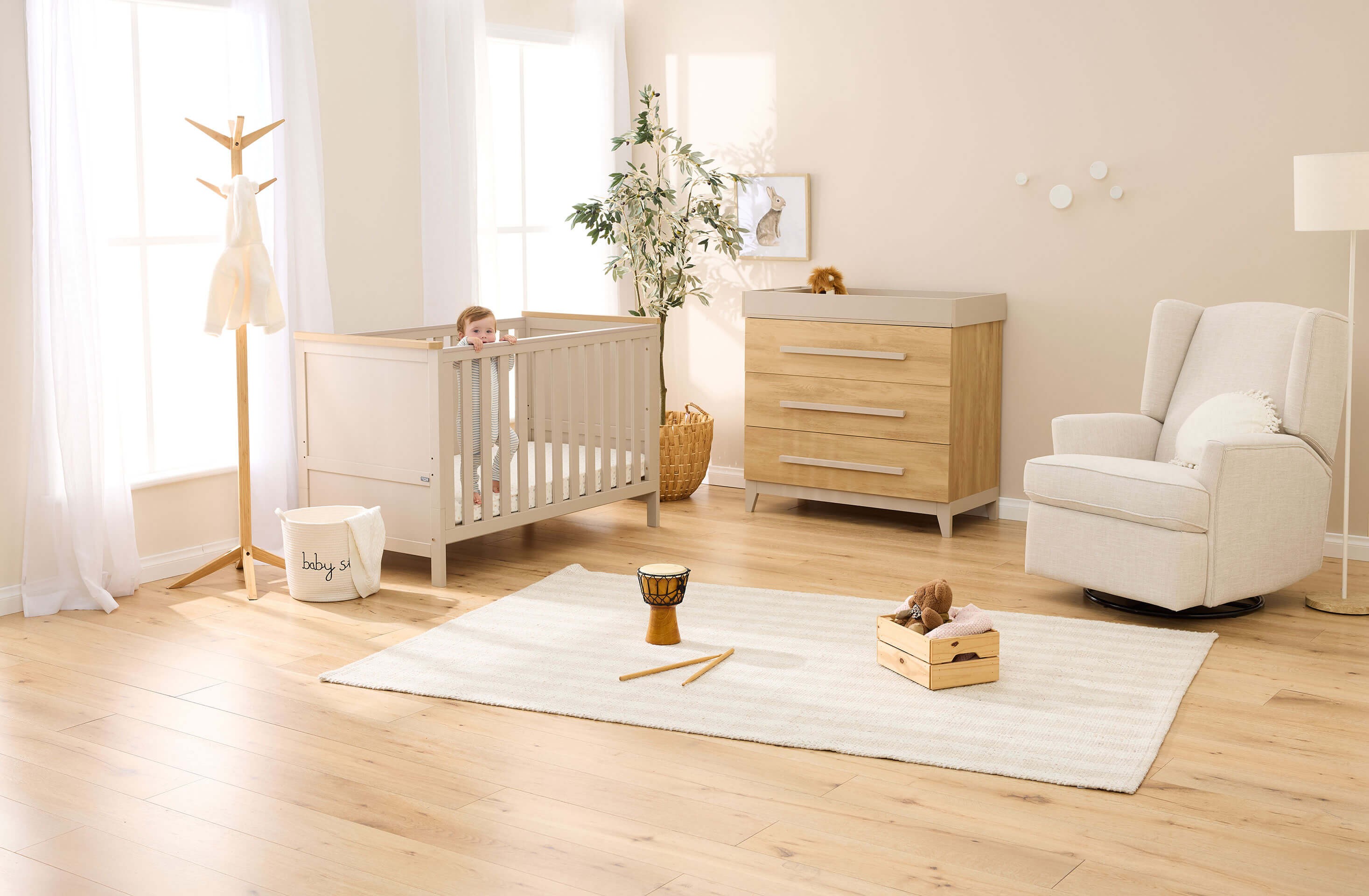 a baby standing in a wooden baby cot inside a nursery room with a matching wooden dresser and a nursing chair and baby playmat