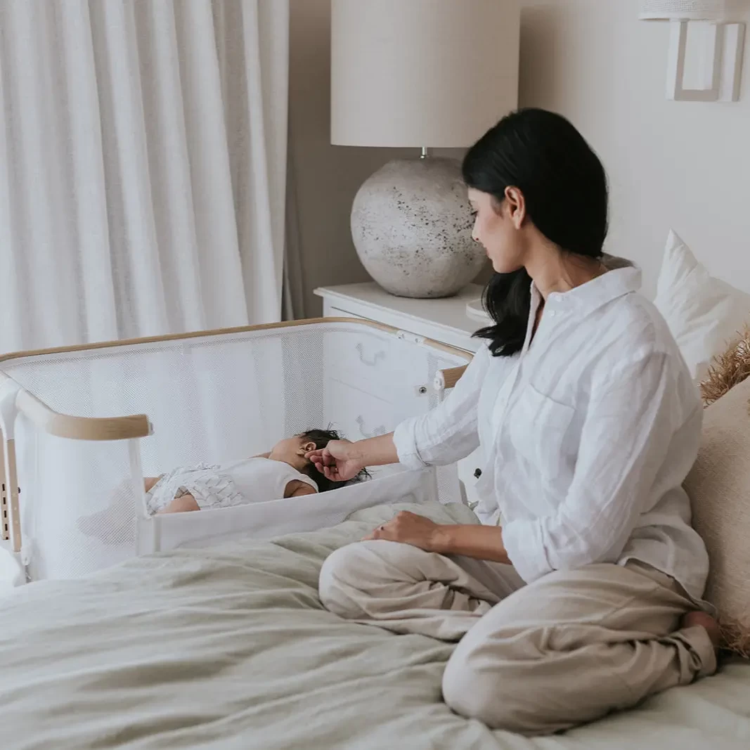 The Babyrest Alfie2 co sleeper sitting next to a bed with a mum soothing her baby in it