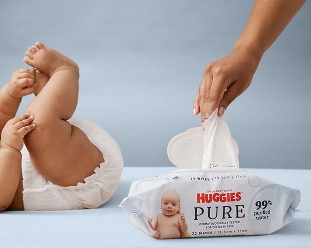 A baby with a nappy on while a mum pulls a Huggies baby wipe out of the packet