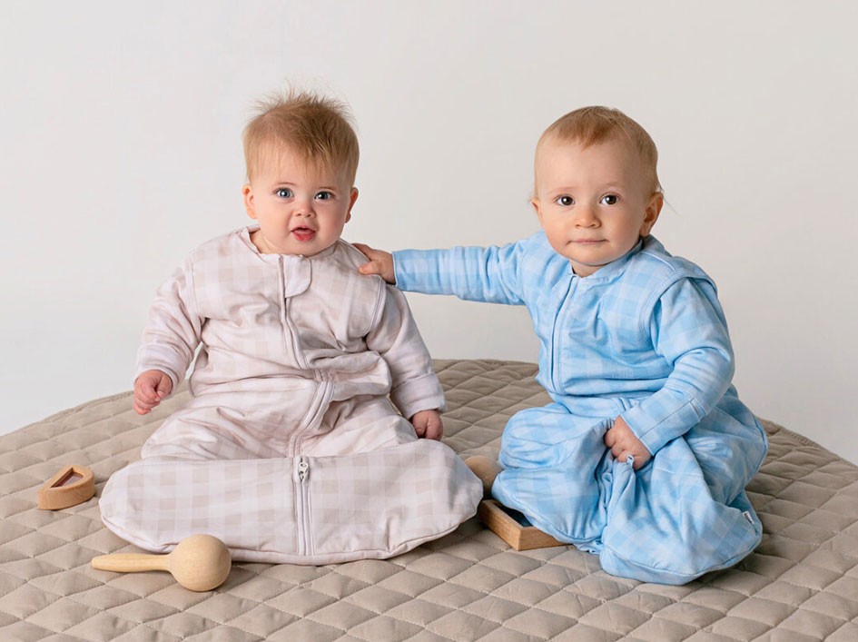 Two babies in Sleepybelly sleeping bags with sleeves playing with wooden toys on a playmat indoors.