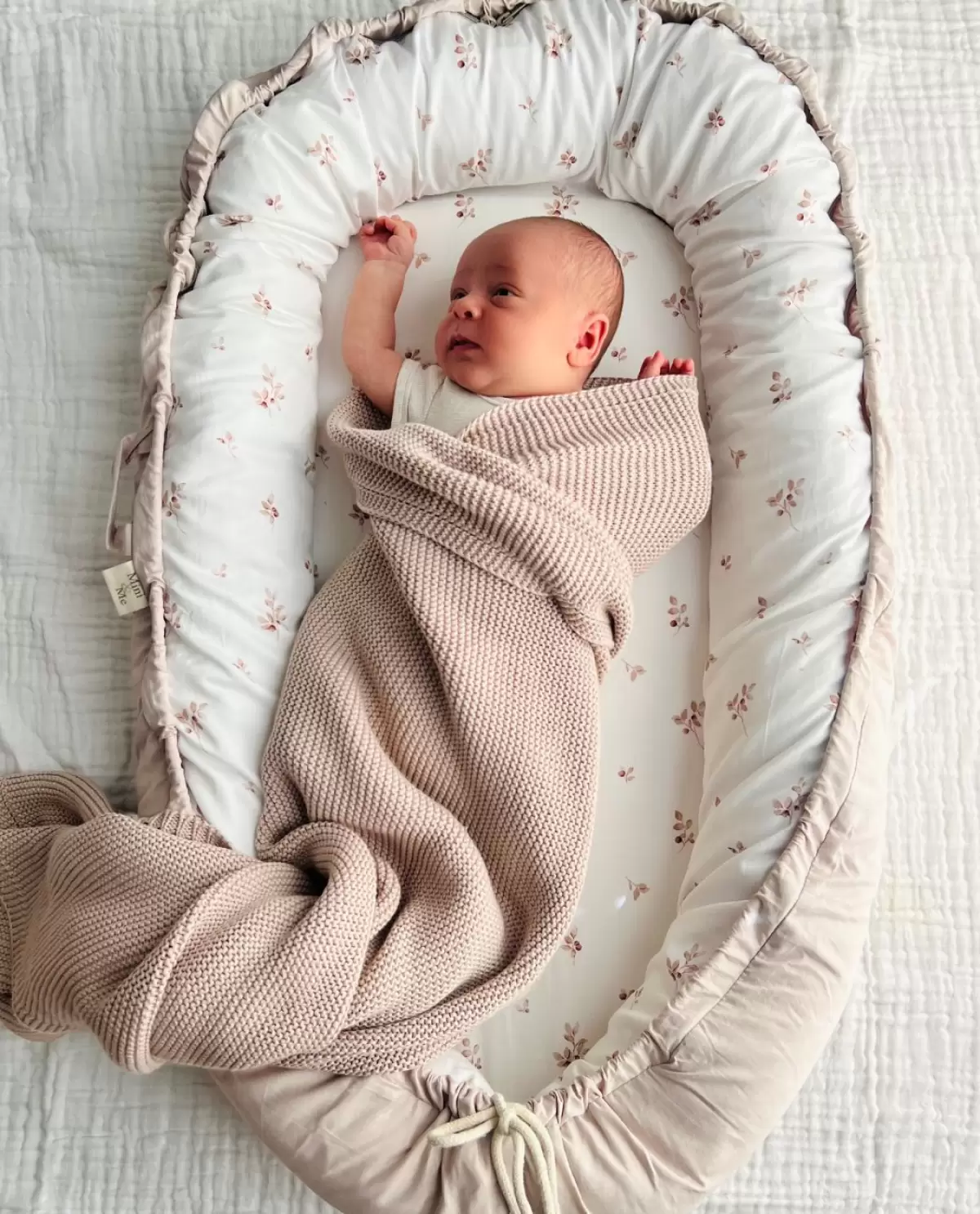 Best infant loungers like this Cuddle Bug & Co fall print baby lounger provide comfort and support – shown with baby wrapped in pink blanket.