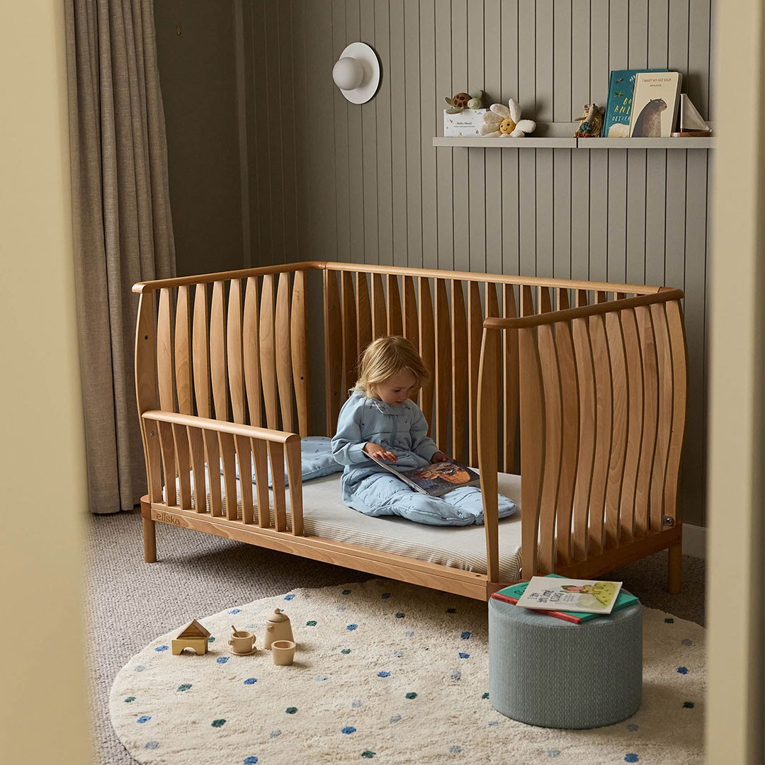 “Wooden baby cot converted to a toddler bed with a child in a sleeping bag reading a childrens book, wooden toys scattered on the bedroom floor.
