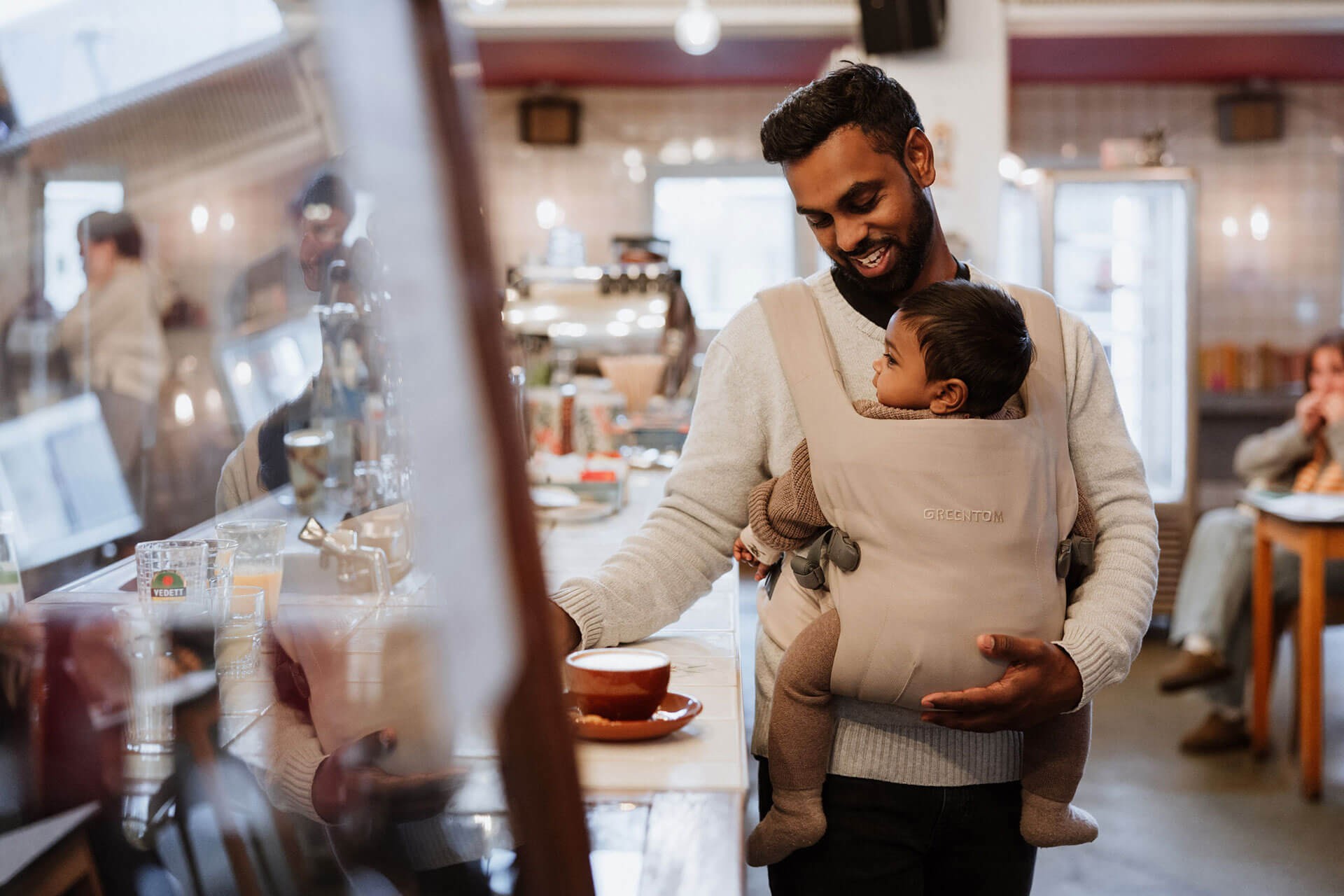 Father carrying baby in the best baby carrier in beige while ordering coffee at a café