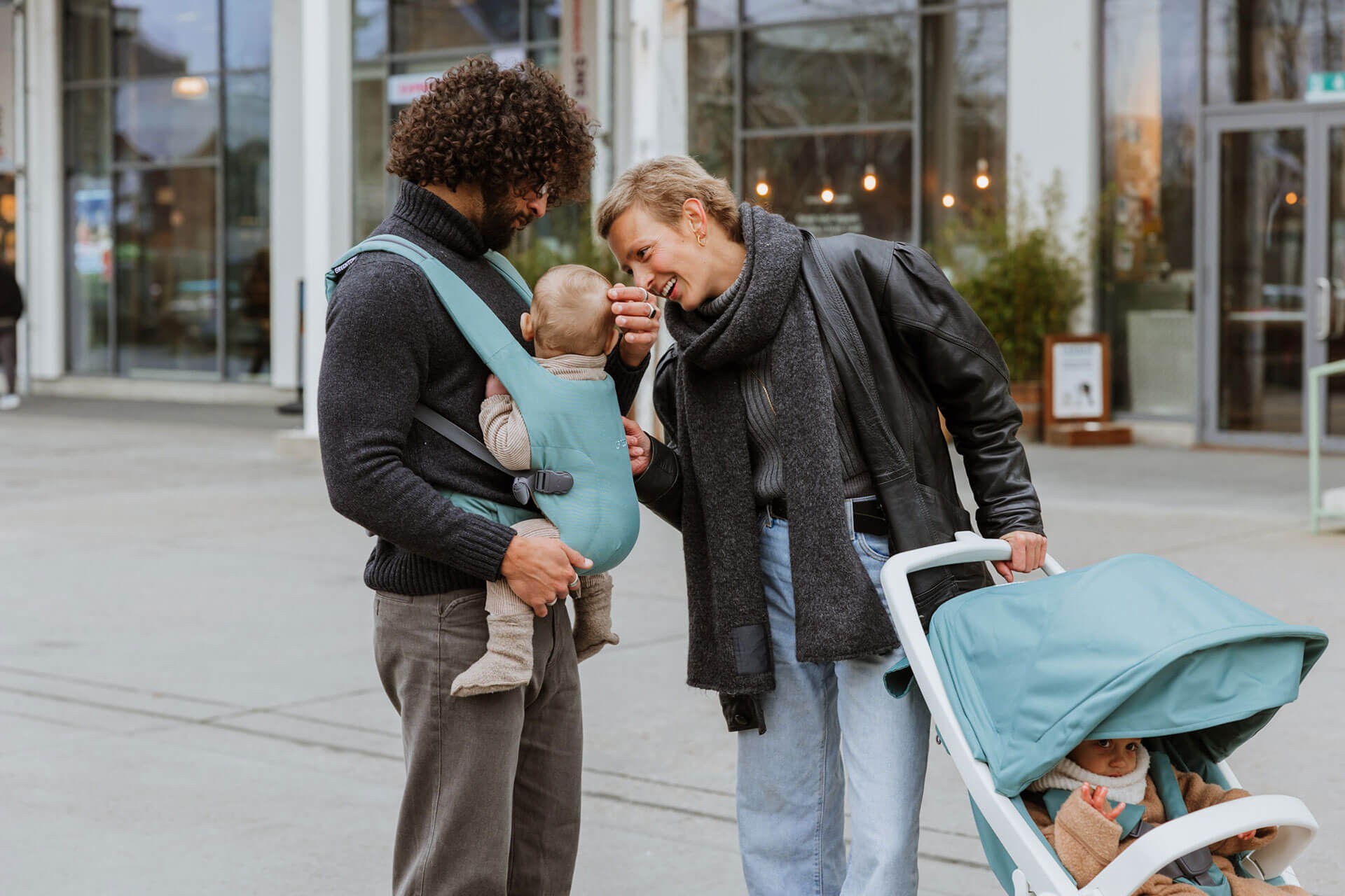 Dad carrying his baby in the best baby carrier in green while mum pushes a toddler in a pram