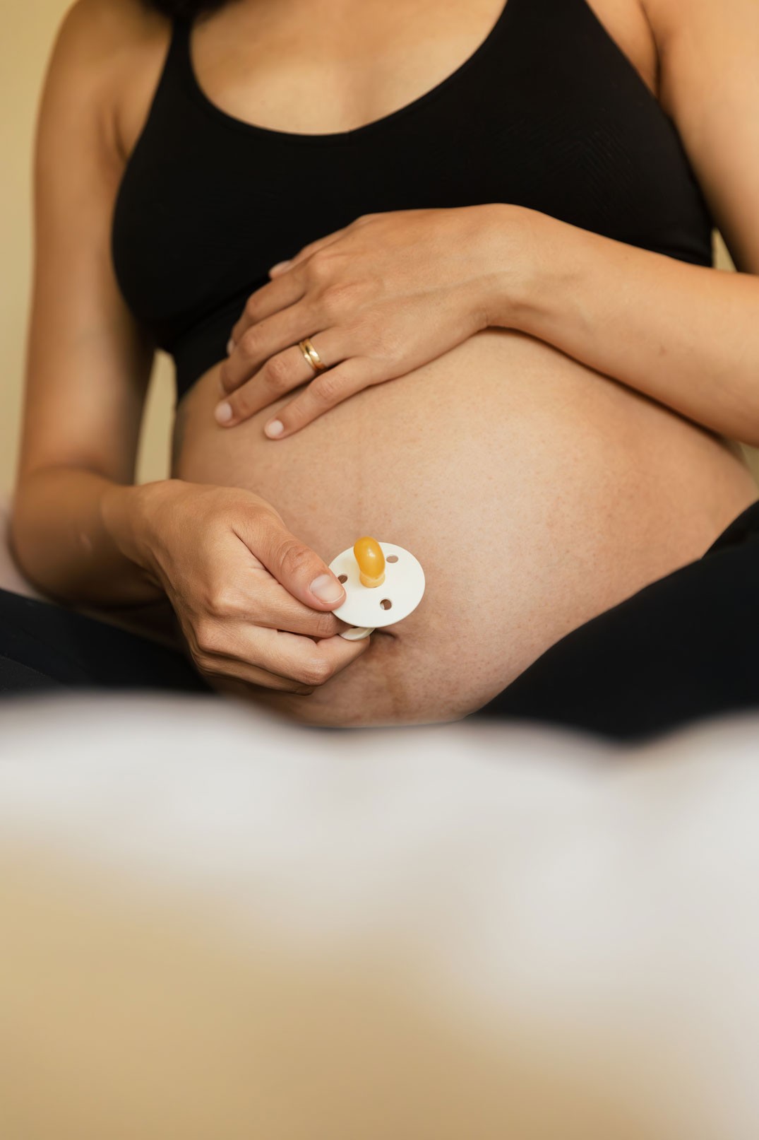 A pregnant woman wearing maternity activewear, seated on the bed while holding her belly bump and a baby dummy by Bib on the other
