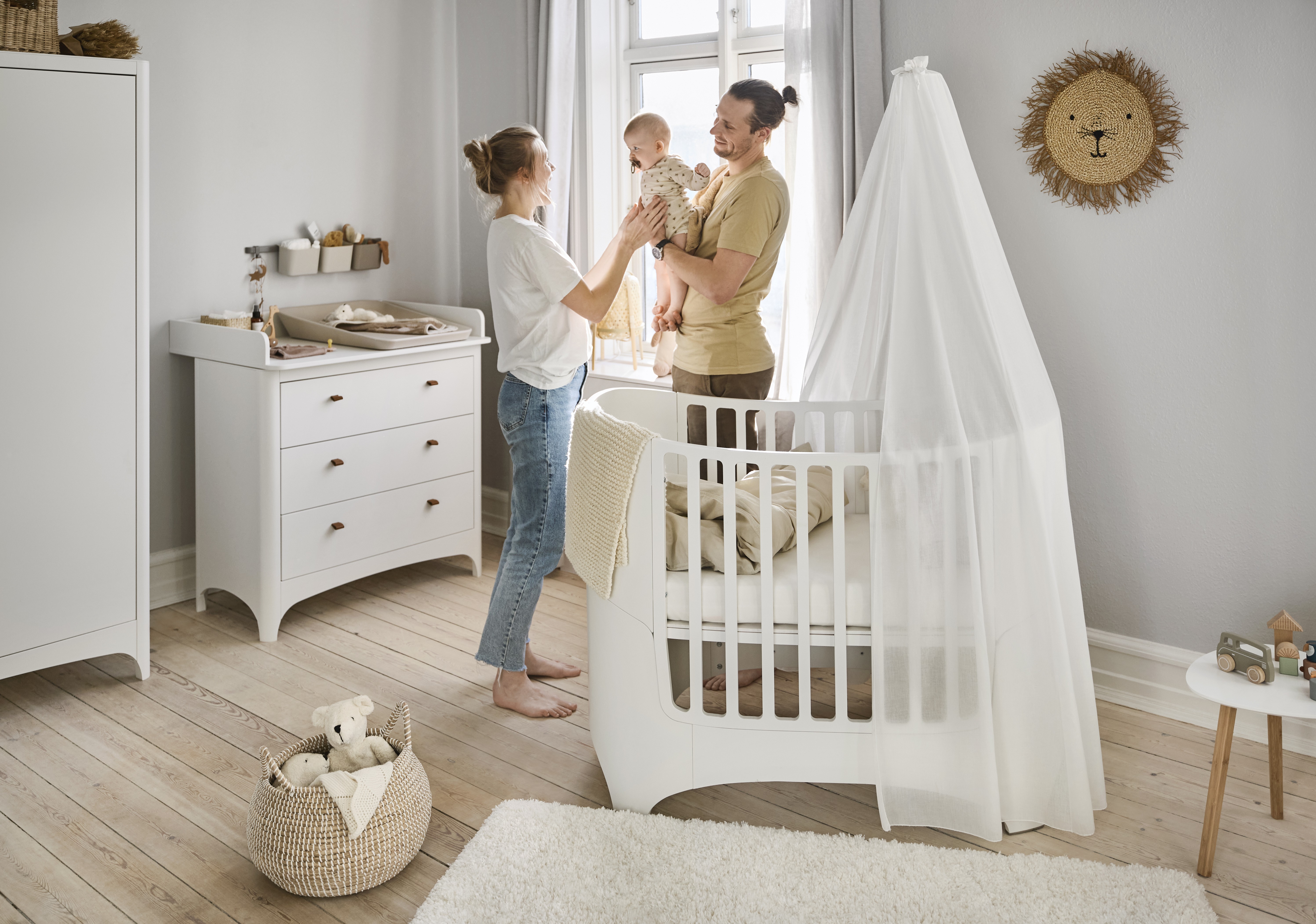 Two mums standing or sitting near a crib; one is holding a baby; the Leander Classic Babycot in whitewash finish is beside them in a nursery setting.