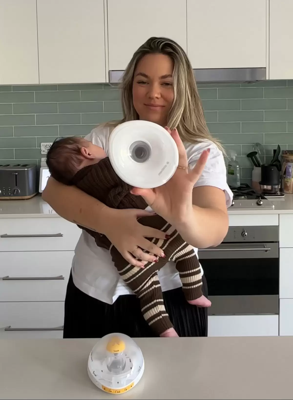 Australian mum holding her baby while showing the Medela Magic InBra hands-free breast pump she uses at home.