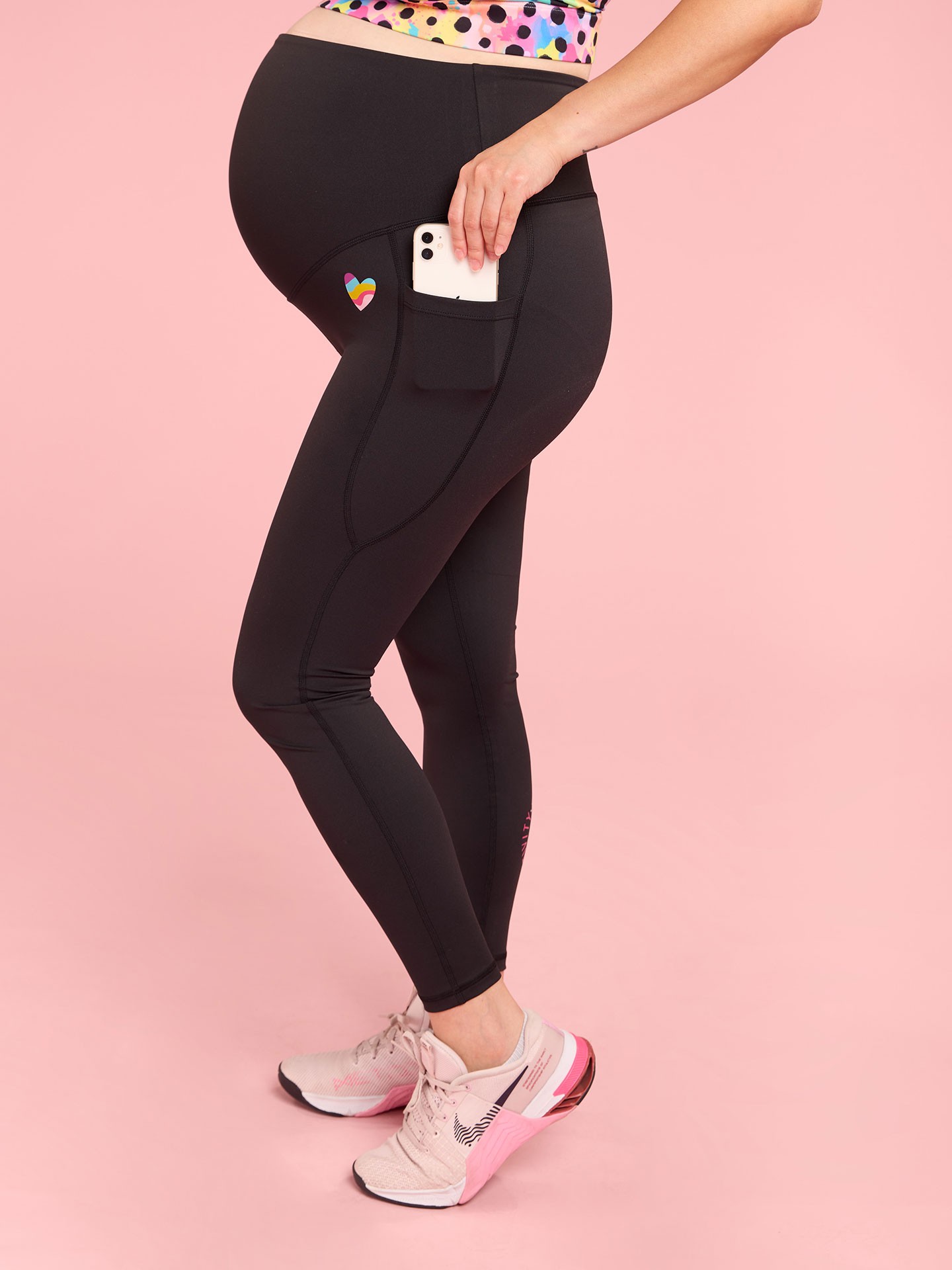 Happy pregnant mum wearing black Mama Movement best maternity activewear leggings with mobile pocket against a light pink background.