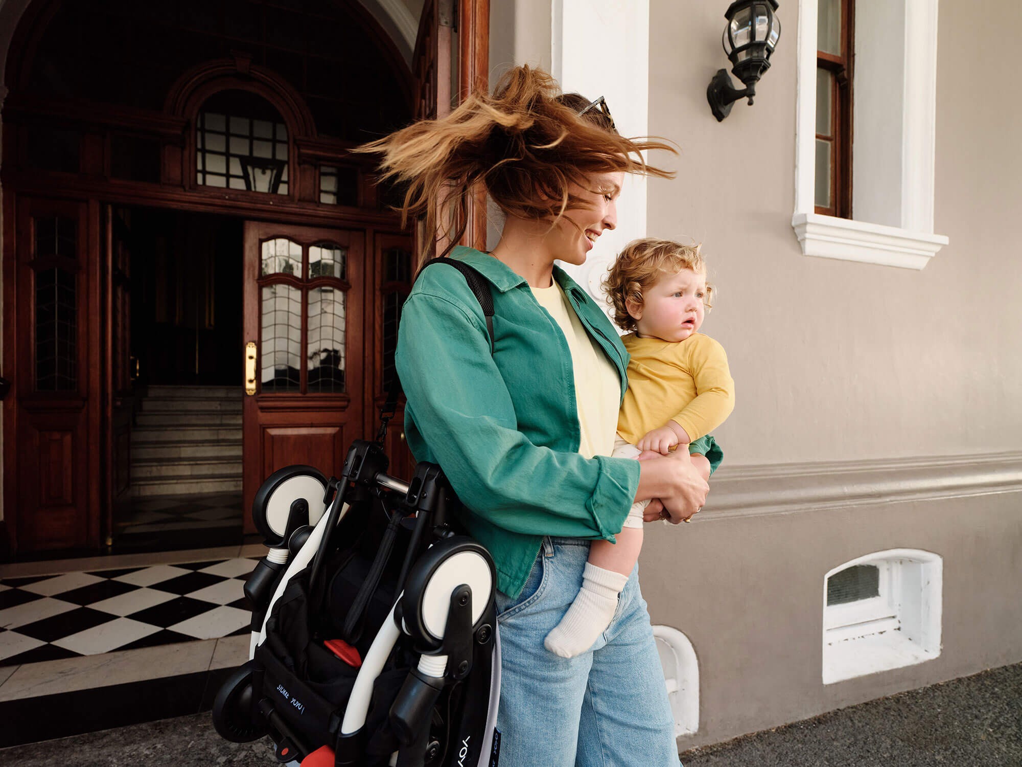 Mum carrying her baby in one arm while holding the folded Stokke Yoyo³ Travel Pram.