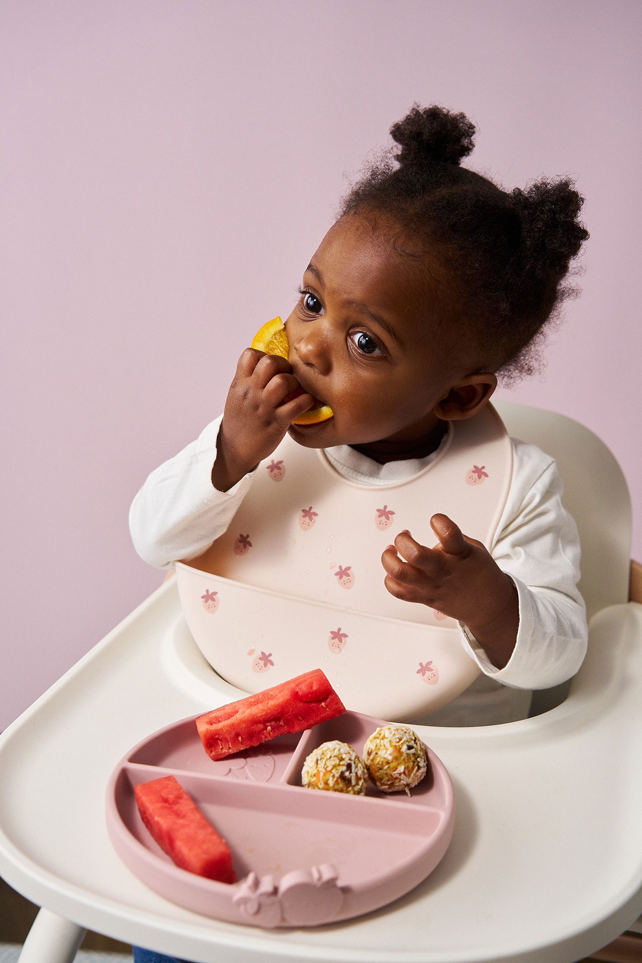 Baby girl seated in a high chair eating watermelon and vegetable balls as she explores first foods for babies at around six months.