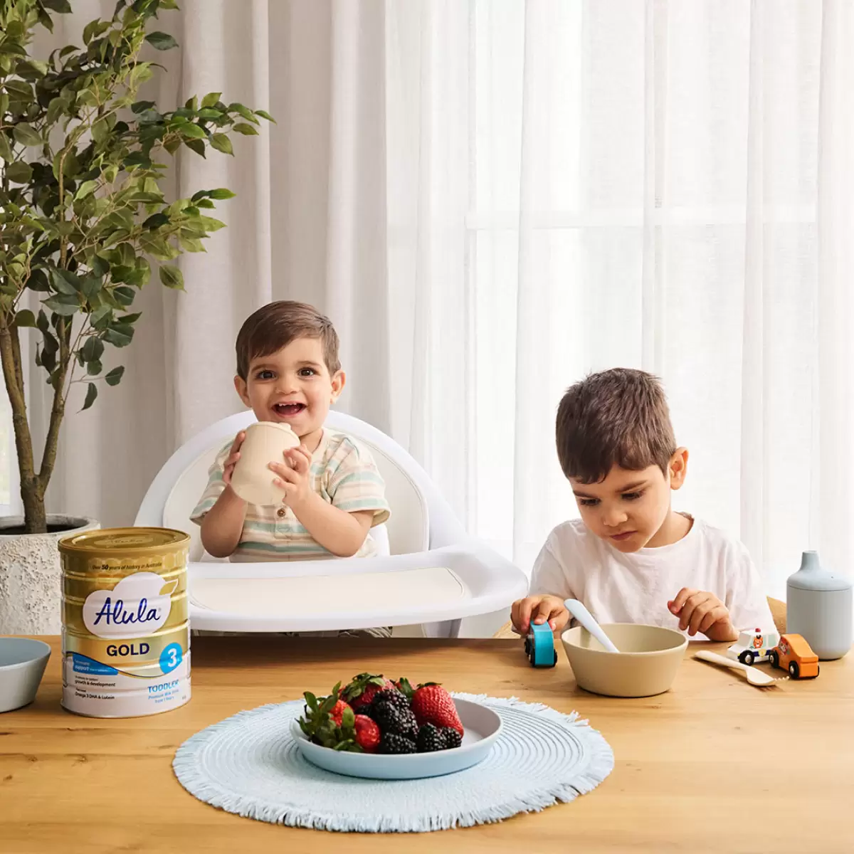 Two children sit at a table, smiling, with a bowl of colorful fruit and a box of Best Formula Milk nearby.