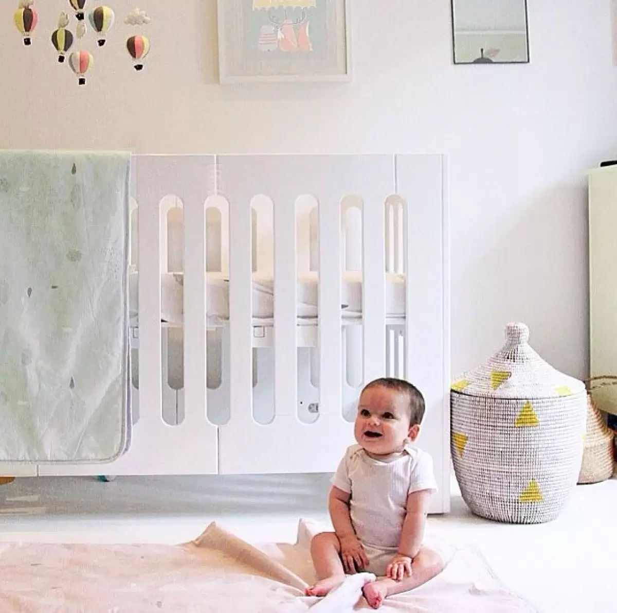 Baby sitting beside the Bloom Alma Papa Cot in Coconut White – spacious and stylish white cot for growing families.