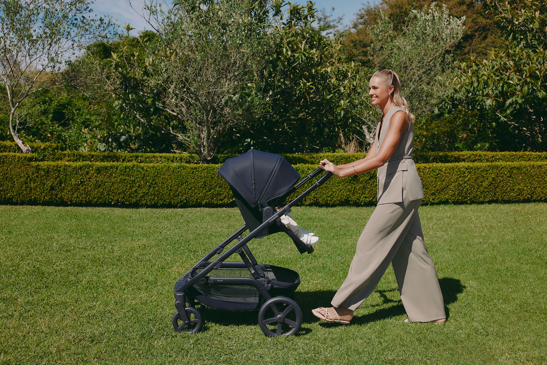 Mum pushing the best single to double pram with the seat facing her and her baby sitting upright during a sunny walk in the park.