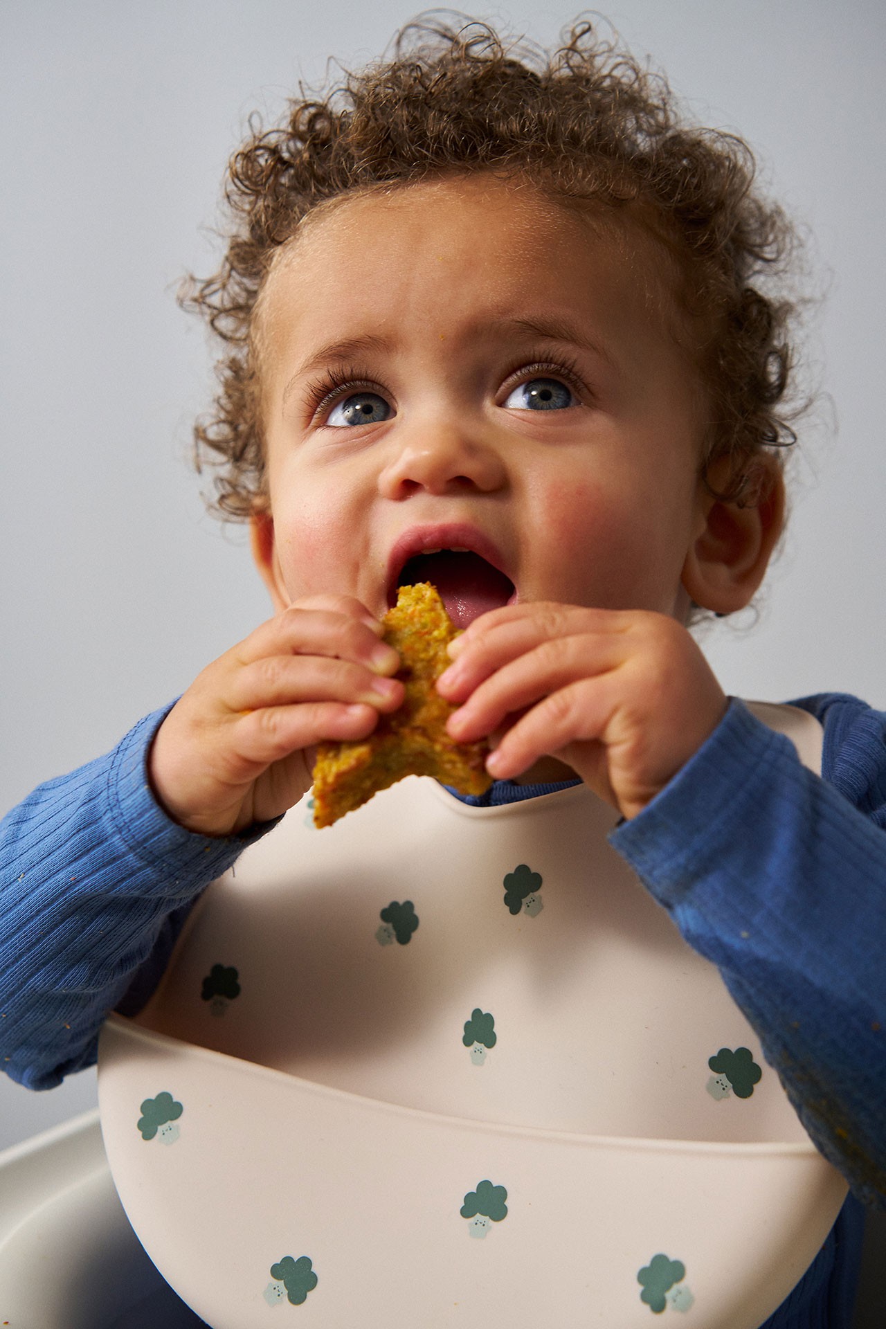 Baby boy enjoying a vegetable fritter as baby led weaning finger foods while seated for mealtime in a silicone bib.