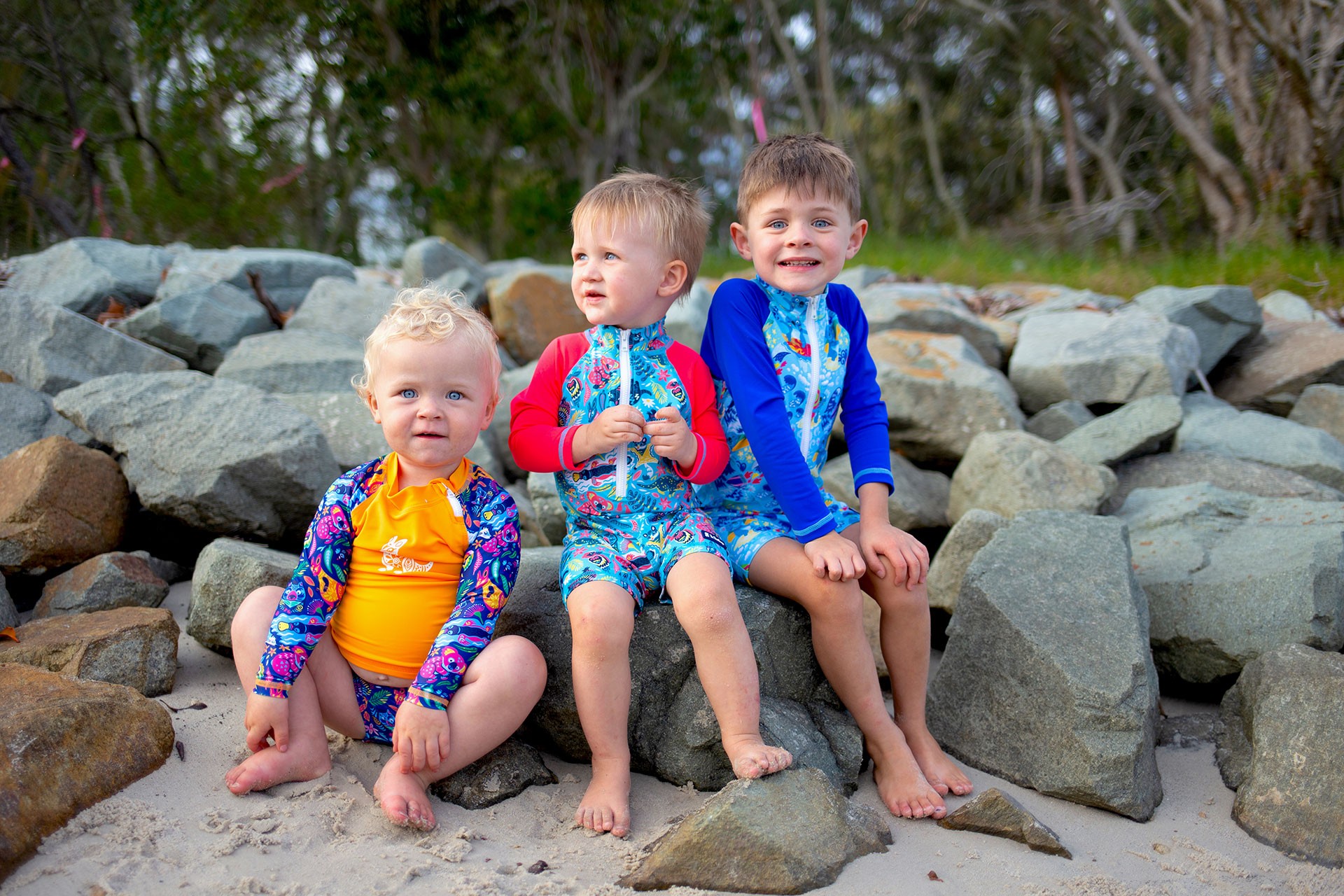 Three kids enjoying the summer in bright kids swimwear in Australia while sitting on a rocky beach.