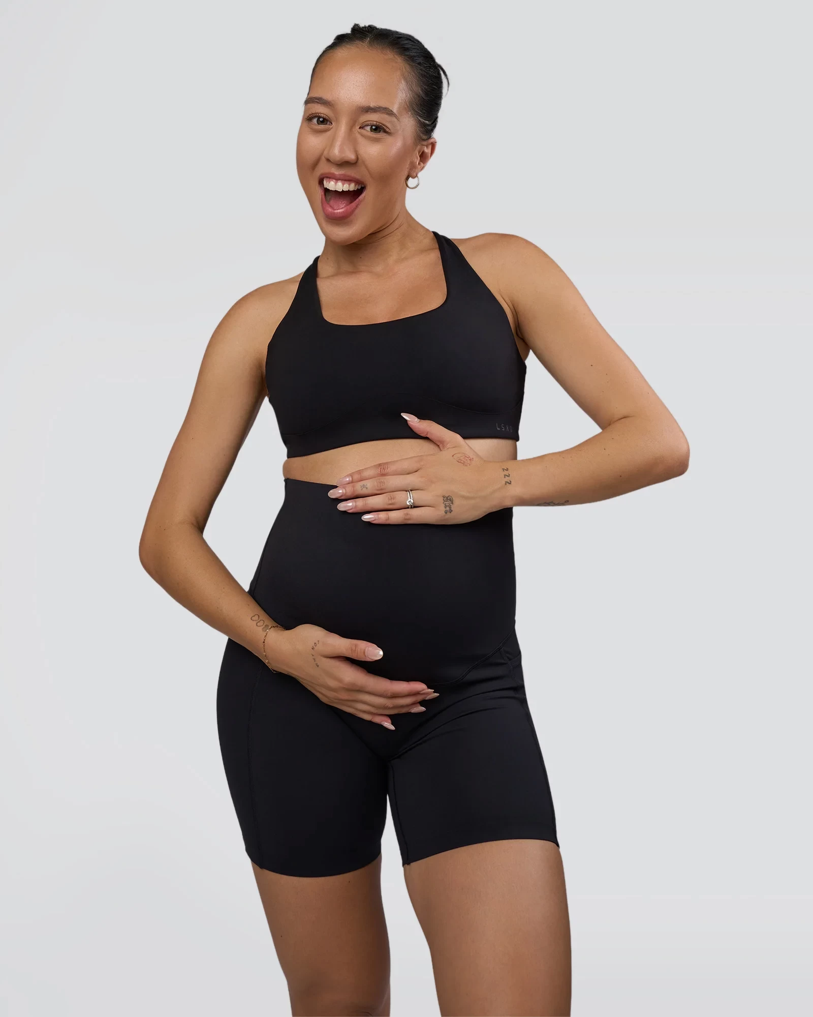 A woman smiling at the camera holding her stomach, wearing maternity activewear bike shorts