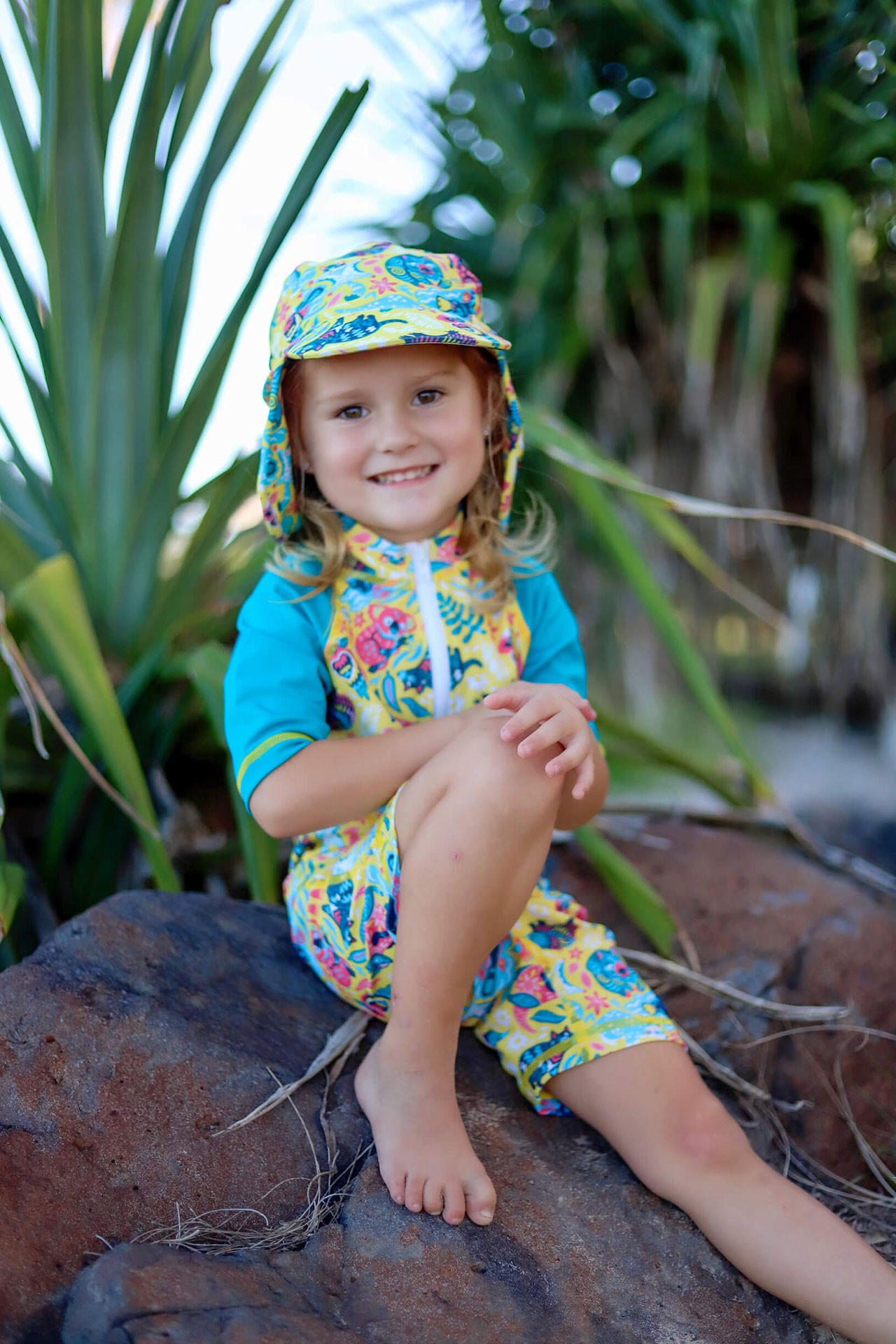 Girl sitting on rocky shoreline smiling in bright printed best kids swimwear in Australia and matching flap hat