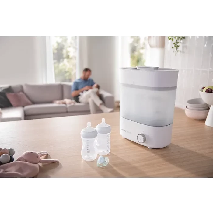 The Philips Avent Steam Steriliser shown on a table with bottles, with a dad in the background feeding a baby