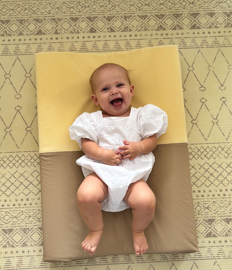 Baby girl in a white romper lying happily on a yellow brown change mat during nappy change time.