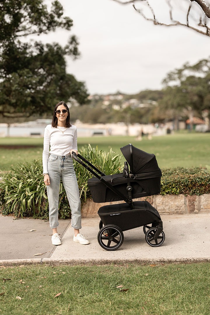 A woman in sunglasses and jeans pushes a baby stroller along a path.