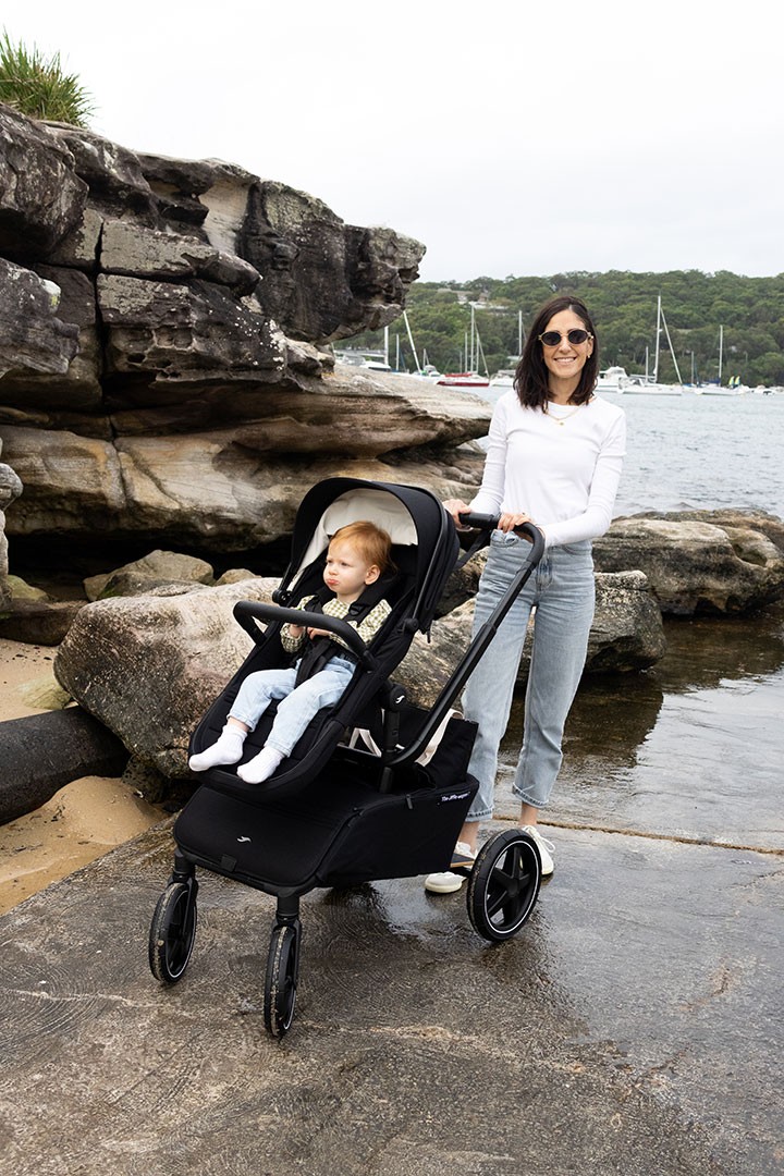 A woman in jeans and sunglasses walks by the shore, pushing a baby in the Best stroller in australia along the water's edge.
