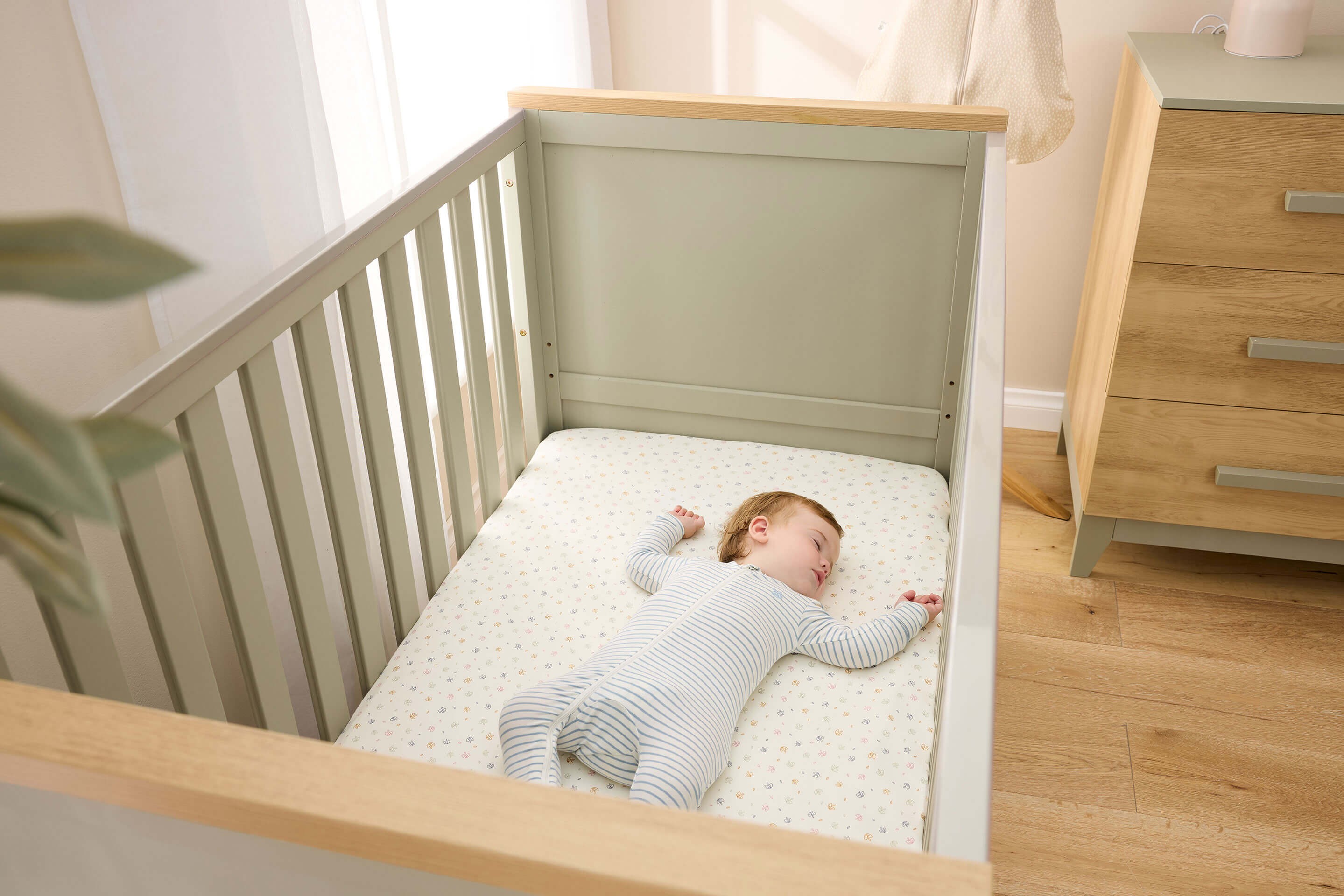 a baby wearing a footed sleepsuit, sleeping on an il tutto misha baby cot in sage colour