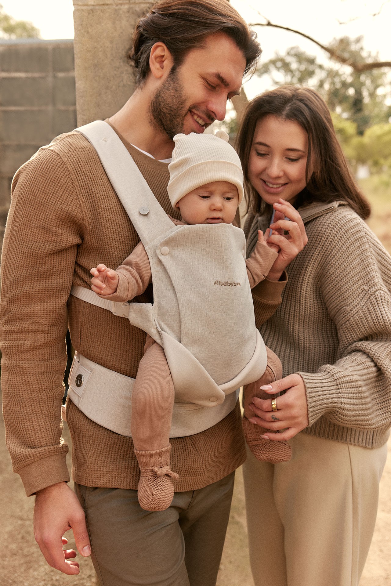 Parents smiling brightly at their child as the dad carries her in a newborn baby carrier during an autumn outing