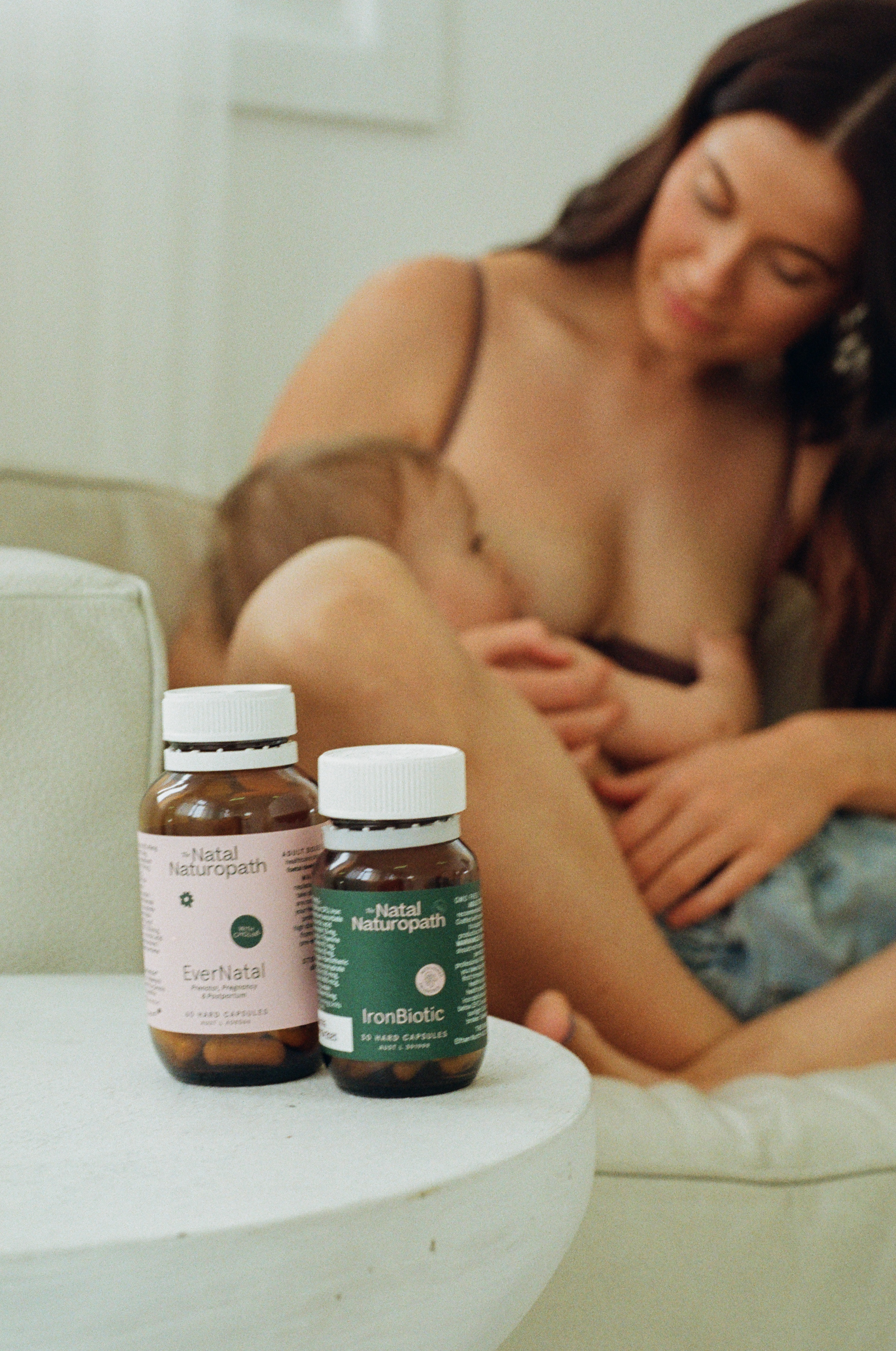 Close-up of two Natal Naturopath EverNatal bottles with mum breastfeeding baby blurred in background