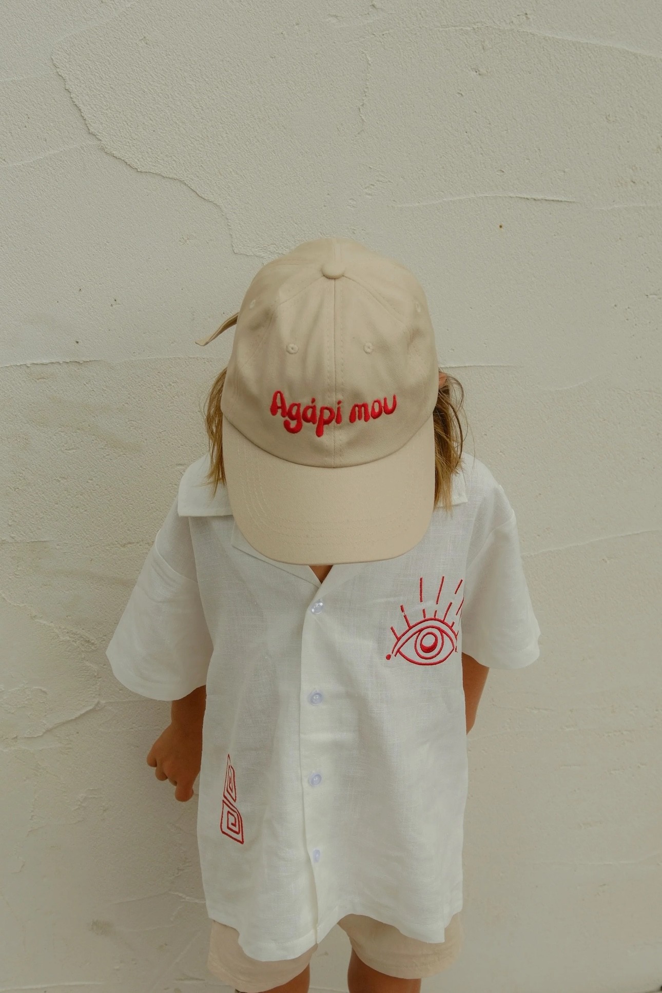 A toddler with his face down to show his hat, that says 'agapi mou', by Nisi Haus children's clothing brand