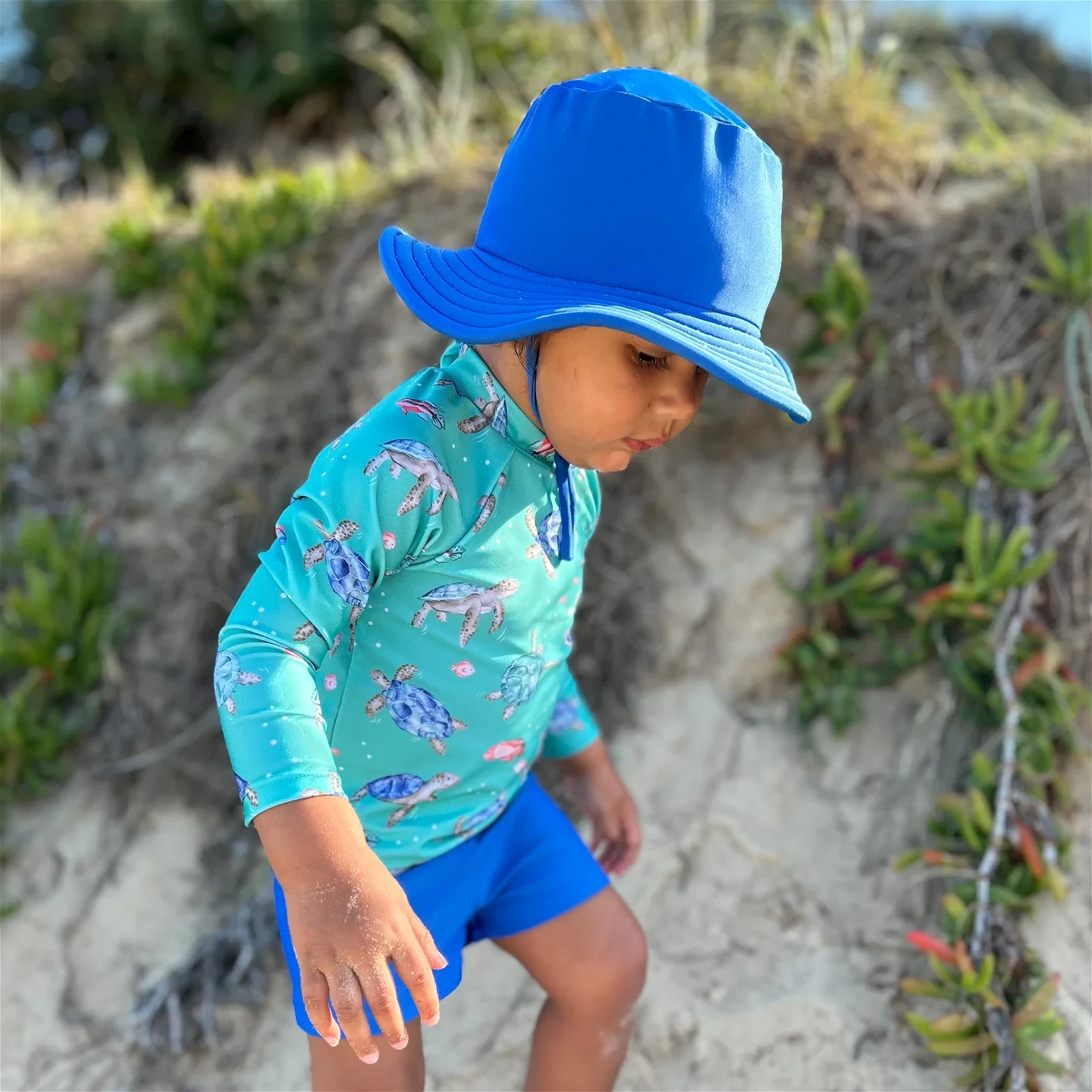 Side profile of toddler boy dressed in purebaby best kids swimwear on beach sand.