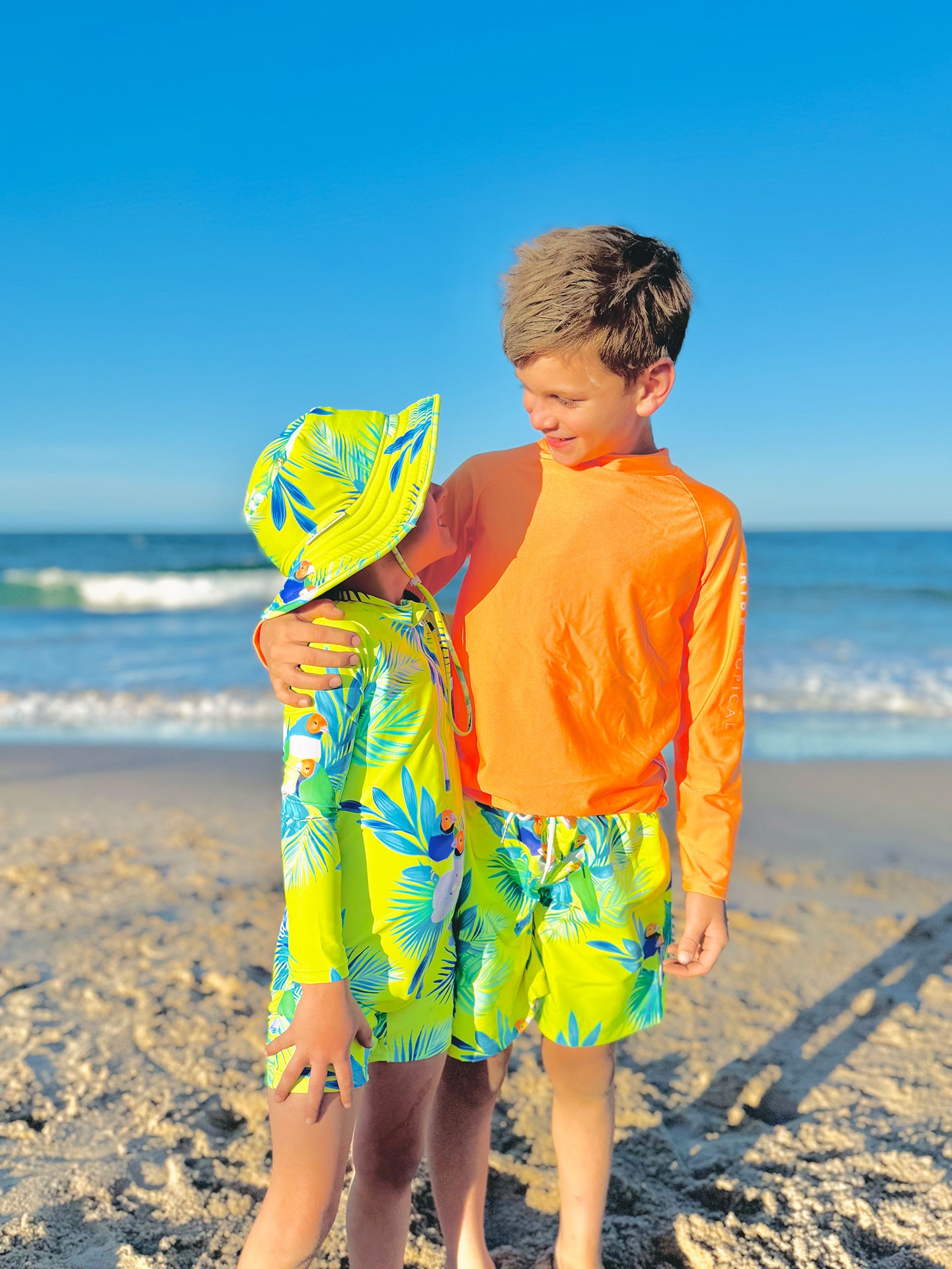 Young boy and toddler wearing bright neon board shorts and rash guards by Tribe Tropical at the beach – best swimwear brand in Australia for kids.